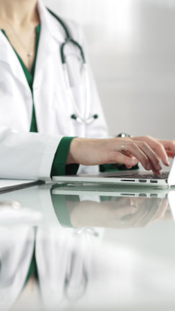 A female doctor in a green blouse under a medical coat is using technology in healthcare. Close-up of hands typing on a laptop on a glass desk in the clinic, vertical view. Medical conceptの写真素材