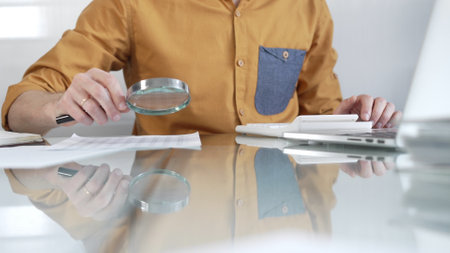 Businessman accountant wearing casual yellow shirt carefully reviewing financial reports with magnifying glass and calculator, conducting audit and analyzing data in office. Taxes, audit in businessの写真素材