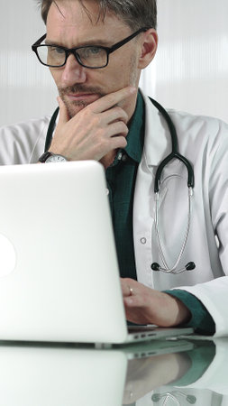 A male doctor in a green shirt under a medical coat is thinking while using technology in healthcare. Man is typing on a laptop on a glass desk in the clinic, vertical portrait view. Medicine conceptの写真素材