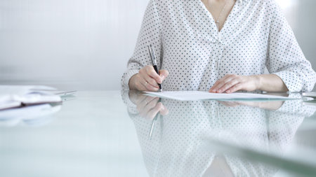 Businesswoman in white dotted blouse is signing a contract at a sleek glass desk in a modern office, showcasing professionalism and focus in her work environment. Business people conceptの写真素材