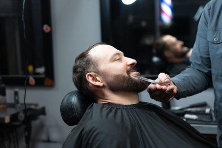 Skilled barber grooming clients beard using professional scissors, delivering precise styling within contemporary barbershop settingの写真素材