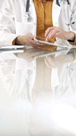Unknown male doctor wearing a stethoscope, yellow shirt under a lab coat, is using digital tablet in modern healthcare setting, interacting with patient data, vertical portrait. Medicine conceptの写真素材