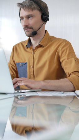 Businessman wearing a headset, focused on a laptop while efficiently working in a modern office environment, vertical portrait. Business people conceptの写真素材