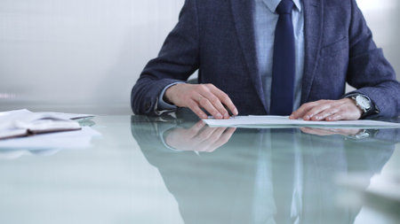 Businessman examining contract terms, using a pen to gesture, in a contemporary office environment, with reflection on a glass desk. Business people or audit conceptの写真素材