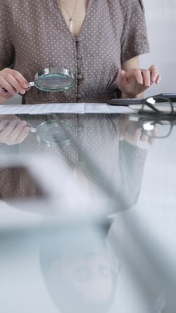 Businesswoman accountant in dotted blouse reviewing financial reports with magnifying glass and calculator in office, vertical view. Taxes, audit in businessの写真素材