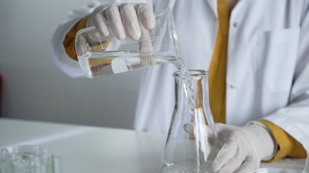 Unknown scientist wearing a lab coat and gloves is carefully pouring a liquid from a beaker into an Erlenmeyer flask, close up. Science and medicineの写真素材
