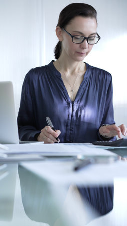 Professional woman accountant in glasses using a calculator and taking notes at a glass desk in the office. Audit and taxes in businessの写真素材