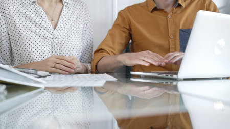 Two unknown accountants are discussing a tax audit, using a laptop and reviewing documents at a glass table, closeup view. Taxes, audit in businessの写真素材
