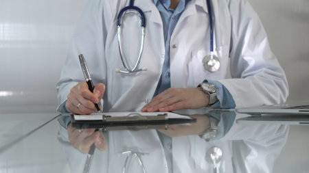 Woman doctor, wearing a blue blouse under a white medical coat, is using a clipboard and taking notes on glass desk in a medical office, close-up. Medicineの写真素材