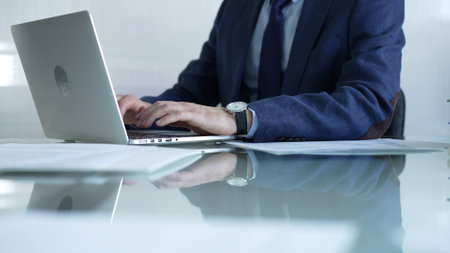Businessman in blue suit is typing on laptop at glass desk while sitting in bright office, showcasing productivity and modern work environment. Businesspeople conceptの写真素材