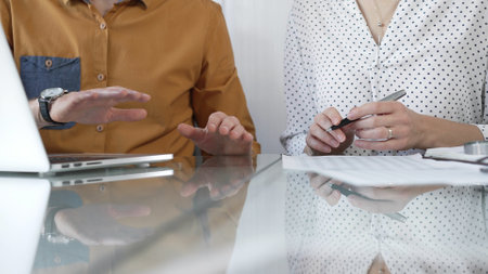 Two professional accountants are discussing a tax audit, using a laptop and reviewing documents at a glass table, closeup view. Taxes, audit in businessの写真素材
