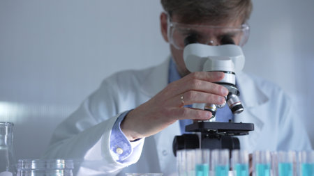 A man scientist, wearing a lab coat, mask, and safety glasses, is using a microscope surrounded by test tubes in laboratory setting. Medicine and science conceptの写真素材