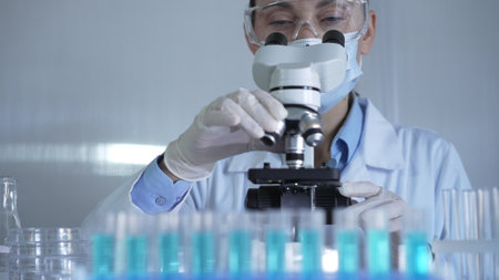 A scientist woman, wearing a lab coat, mask, white gloves and safety glasses, is adjusting a microscope surrounded by test tubes in laboratoryの写真素材