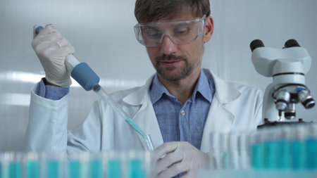 Scientist man wearing a lab coat and safety glasses is using a micropipette to drop a liquid into a test tube, portrait view. Medicine and scienceの写真素材