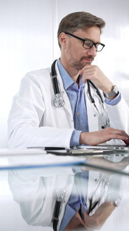 Doctor man in a blue shirt under a medical coat is thinking while using technology in medicine. Portrait of physician typing on a laptop on a glass desk in clinic, vertical view. Medicine and scienceの写真素材