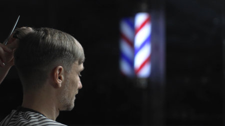 Professional barber grooming male clients hair with scissors, vintage barber pole framing traditional barbershop settingの写真素材