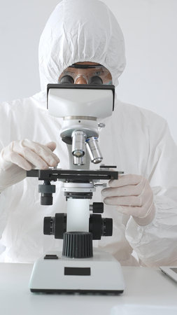 Medical virologist in protective hazmat gear examining viral samples under a microscope while...