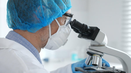 Male scientist wearing protective gear examining microscope slide, analyzing research sample in...