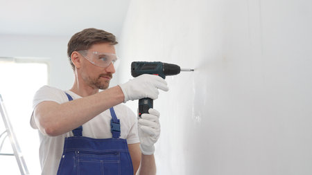 Male construction worker in a blue jumpsuit, safety goggles, and work gloves is screwing a screw...