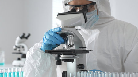 A medical virologist in protective gear and blue gloves examines viral samples under a microscope...