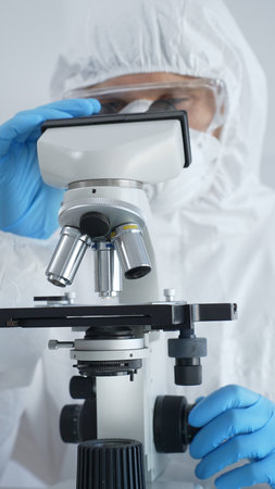 Male virologist in a hazmat suit, mask, and blue gloves, diligently working at a microscope in a...