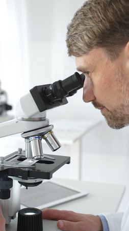A man scientist carefully observing through a microscope in a brightly lit laboratory, conducting...