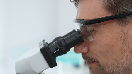 Mature scientist examining specimen under microscope, wearing protective eyewear in contemporary...
