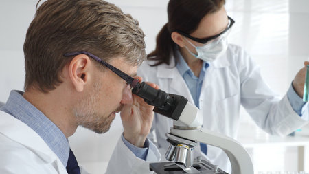 A researcher wearing glasses examines a sample under a microscope, a lab technician wearing a...