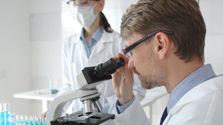 A male researcher examines a sample under a microscope, a female lab technician in a protective mask...