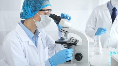 Woman scientist wearing blue protective gear is examining specimen under microscope while laboratory...