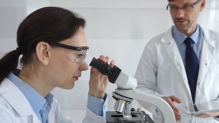 A female scientist wearing protective glasses looks into a microscope and conducts a medical...