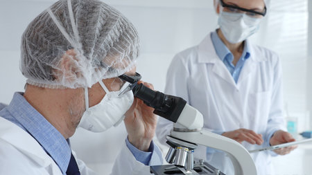 Male scientist wearing protective mask, glasses and hairnet working with microscope while female colleague helping him holding tablet and taking notes in laboratory. Medicine and science conceptの写真素材