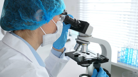 Female scientist wearing a mask and blue safety gear, using a microscope for research while a male...