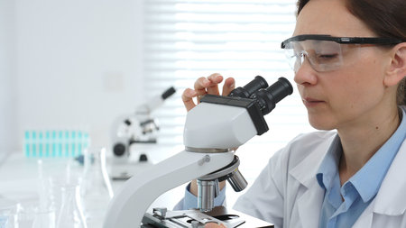 Female scientist examining samples through a microscope in a modern lab, conducting pharmaceutical...