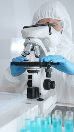 Scientific researcher wearing protective gear adjusting microscope, analyzing lab samples with...