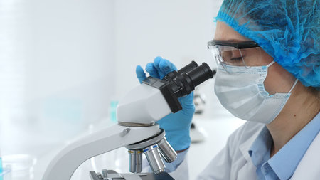 Female scientist analyzing sample through microscope, wearing protective lab gear during medical...