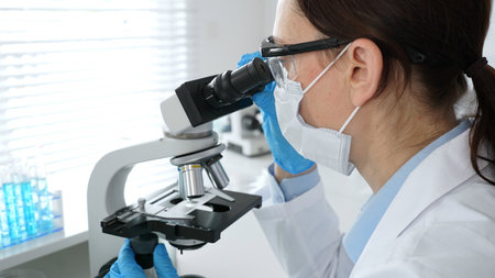 A female scientist in a blue mask and gloves is using a microscope and conducting scientific...