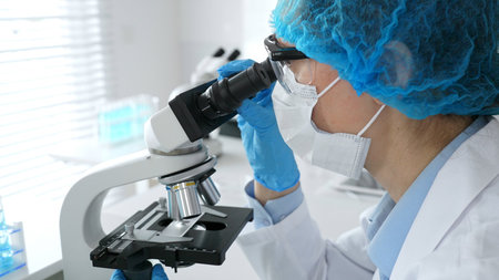 Female scientist wearing a mask and blue safety gear, using a microscope for research while a male...