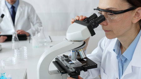 Female scientist examining sample under microscope, conducting research in laboratory with colleague...