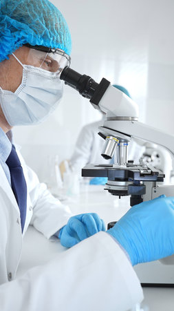 Senior male scientist wearing a protective mask, cap, and gloves, examining samples under a...