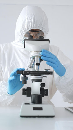 Medical scientist wearing protective suit using microscope while analyzing samples, conducting...