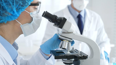 Close up of woman scientist wearing blue protective gear while examining specimen under microscope,...