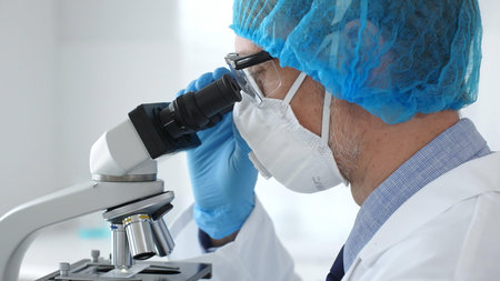 Male scientist wearing protective gear examining microscope slide, analyzing research sample in...