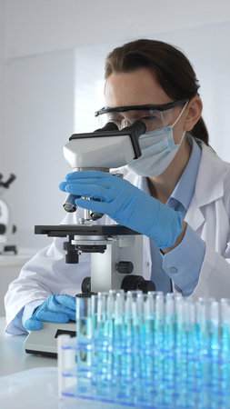 Female scientist examining microscope slide, wearing protective gear in laboratory, researching...