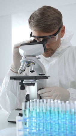 Male virologist in medical gown and gloves examines virus samples under microscope while conducting research in sterile laboratory conditions. Close-up of test tubes with blue liquidの写真素材