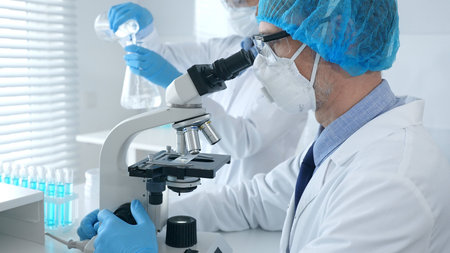 Masked male scientist examining specimen under microscope while laboratory assistant handles...