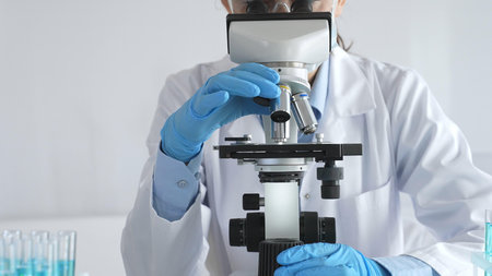 A female scientist in protective gear adjusts a microscope lens and accurately analyzes medical...