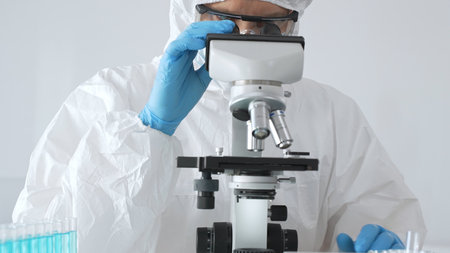 A male doctor in a protective suit and gloves uses a microscope while analyzing samples, conducting...