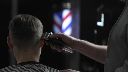 Male professional barber trimming clients hair with electric clipper, vintage illuminated barber pole visible in background, creating contemporary grooming experienceの写真素材