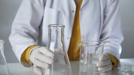 Unknown scientist wearing a lab coat and gloves is carefully pouring a liquid from one beaker into...